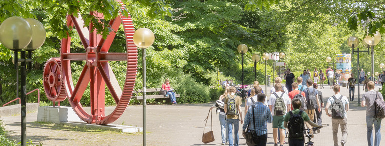 Students walking across Campus Nord. In The background there is a red sculpture of large gears.